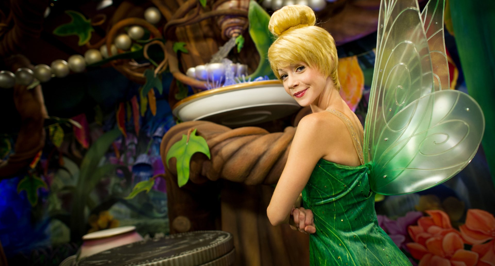 An actress dressed as Disney's Tinkerbell standing in foreground with arms crossed, forest in background..